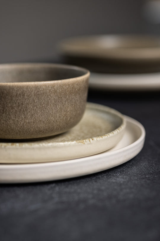 Serviesset in de natuurlijke kleuren bruin, beige en wit, op elkaar gestapeld op een donkerkleurige tafel. Perfect voor een Japandi interieur.