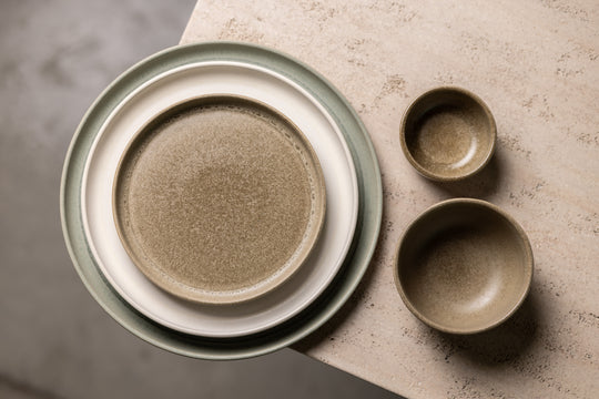 Opgestapelde driekleurig serviesset van Mimo Homeware, er naast twee bruine kommetjes op een natuurstenen tafel.