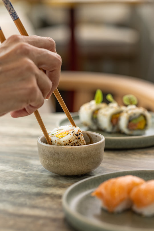 Mini kommetje van bruin Portugees keramiek van Mimo Homeware met sojasaus, waarin sushi wordt gedipt met eetstokjes.